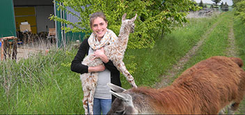 Eine Frau steht auf einem Feldweg in der Eifel und lächelt in die Kamera. Neben ihr steht ein Lama und sie hält ein kleines Lama auf dem Arm.